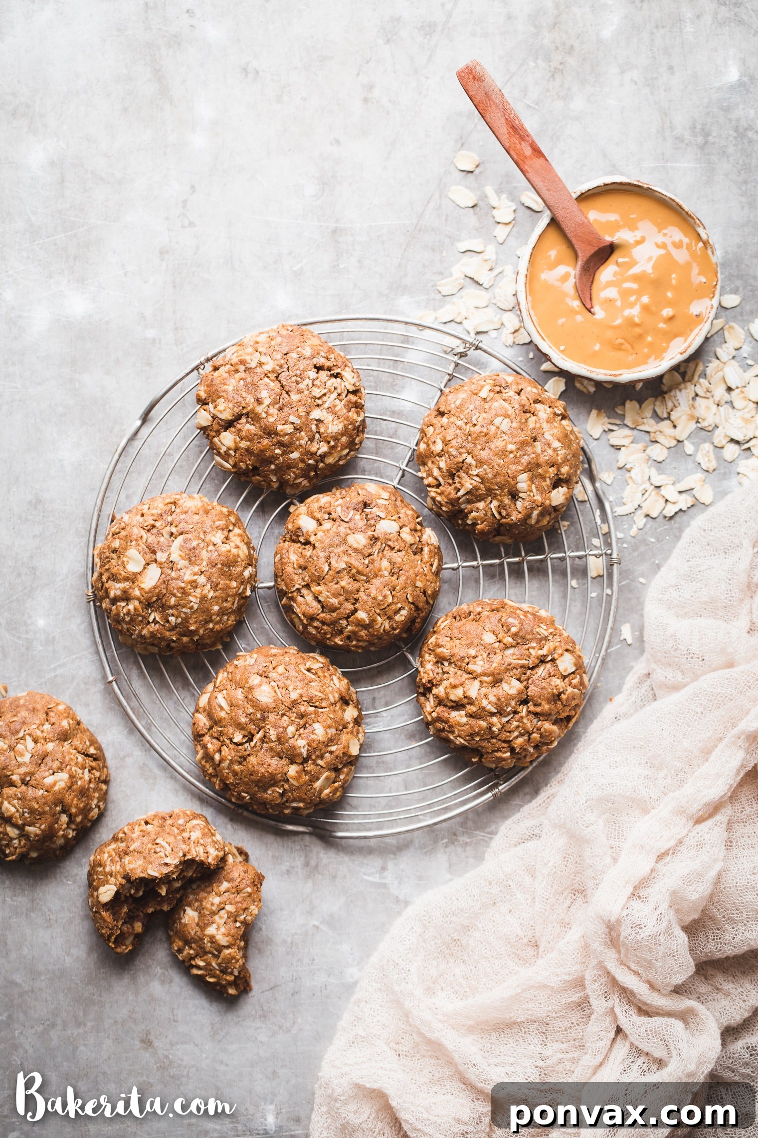 Six perfectly baked Peanut Butter Oatmeal Cookies arranged on a small round cooling rack, set atop a silver baking sheet. A small bowl of creamy peanut butter and a scattering of rolled oats are visible in the background, highlighting the key ingredients.