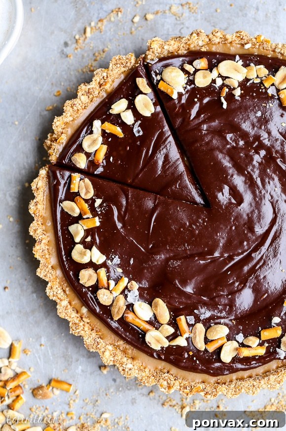 A close-up of a slice of Vegan Chocolate Peanut Butter Tart, showcasing its distinct layers of crunchy pretzel crust, creamy peanut butter filling, and smooth chocolate ganache, garnished with pretzels and peanuts.