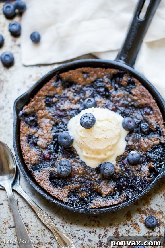 Gooey Blueberry Skillet Cookie served in a cast iron pan, topped with melting vanilla ice cream, showcasing fresh blueberries and a golden-brown crust. This recipe is gluten-free, Paleo, and refined sugar-free.
