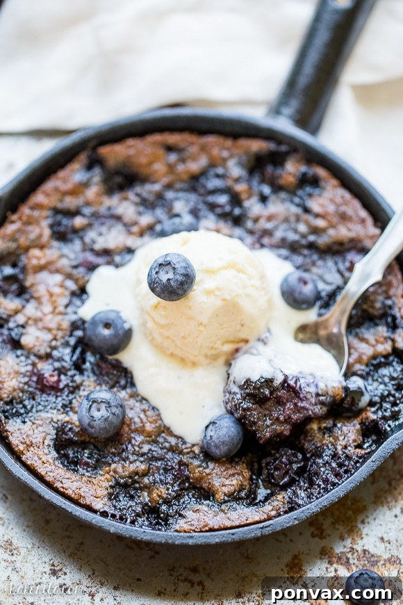 An overhead shot of a Blueberry Skillet Cookie, still in the pan, with a spoon digging into the soft, fruit-filled center, showing the irresistible texture.