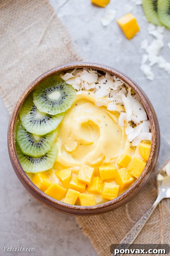 Tropical Mango Pineapple Smoothie Bowl 4 A close-up shot of a Mango Pineapple Smoothie Bowl adorned with vibrant kiwi slices, chunks of fresh mango, and delicate coconut flakes, served in a wooden bowl with a spoon resting on a sheet pan.