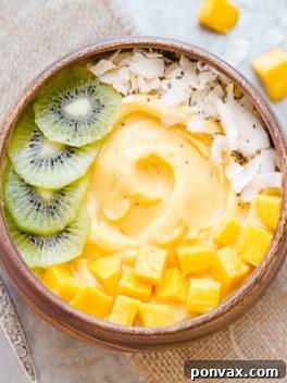 Kiwi slices, mango chunks, chia seeds, and coconut flakes topping a Mango Pineapple Smoothie Bowl in a wooden bowl with a silver spoon, presented beautifully.