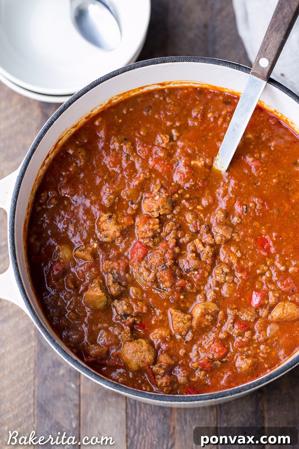 Close-up of a spoon scooping a portion of rich, red Paleo Chili, highlighting the chunks of beef, sausage, and peppers. The deep color and thick consistency are visible, with a sprinkle of fresh green herbs.