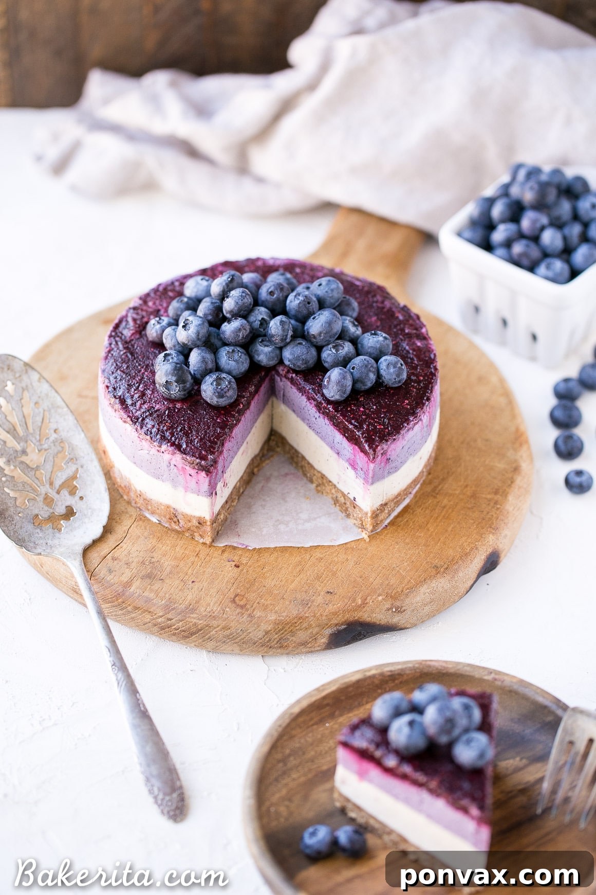 A stunning slice of no-bake layered blueberry vegan cheesecake, showing distinct creamy white and purple layers, with a fresh blueberry topping, on a clean white surface.
