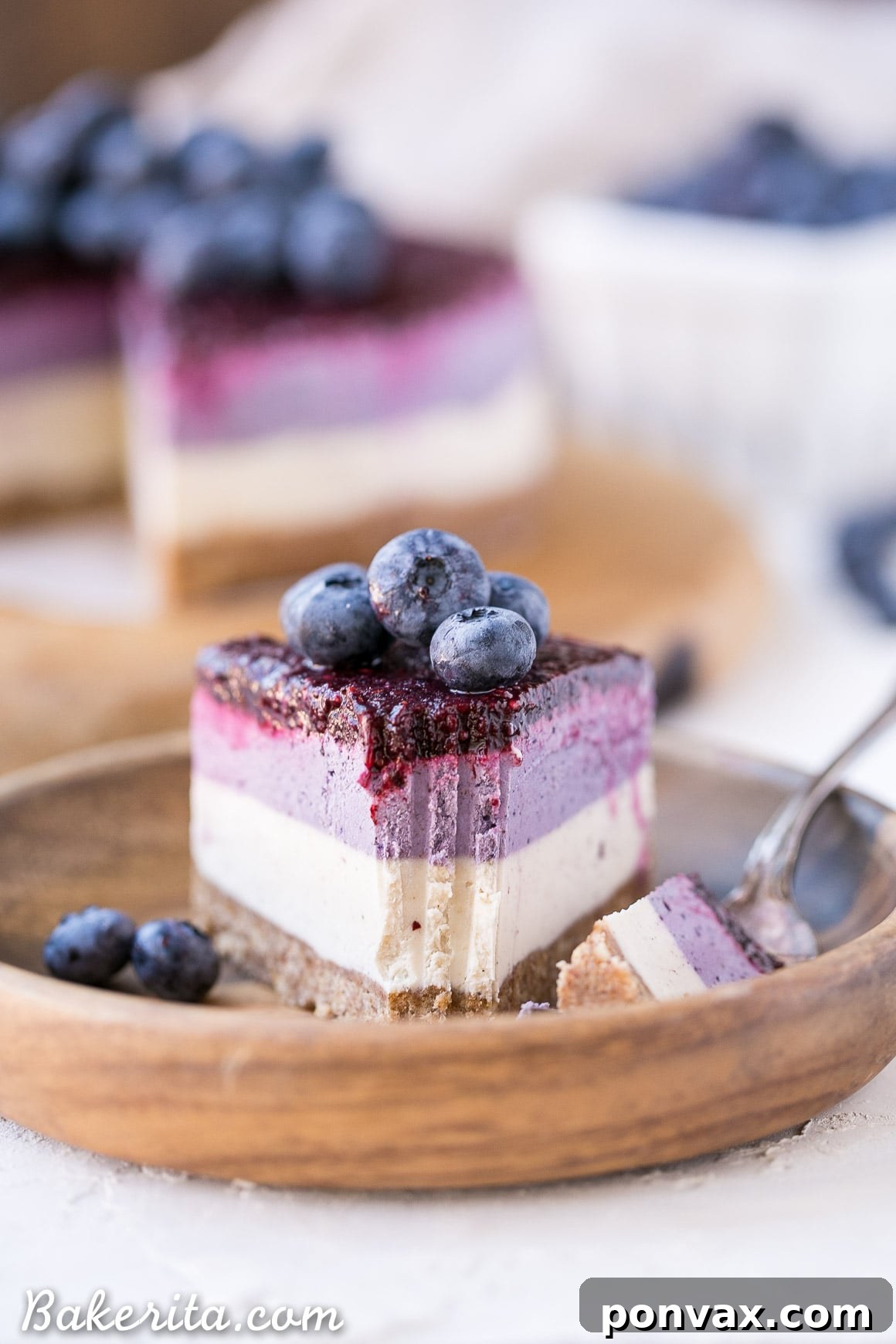 A top-down view of a partially sliced no-bake layered blueberry vegan cheesecake, showing the distinct white and purple layers, garnished with fresh blueberries.