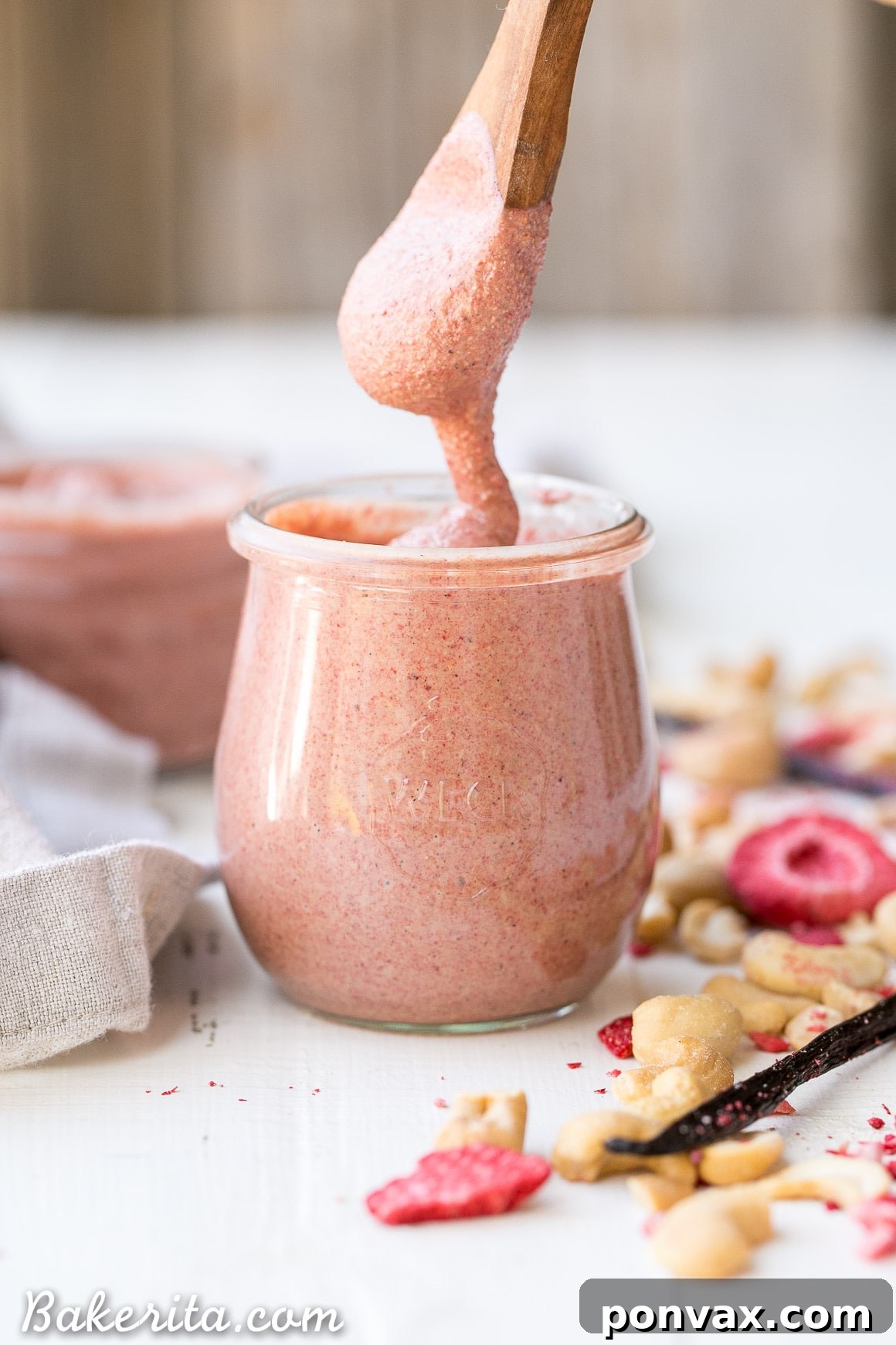 A jar of homemade Strawberry Cashew Butter, garnished with fresh strawberries and vanilla bean. The butter is a beautiful pink color, showcasing its natural ingredients.