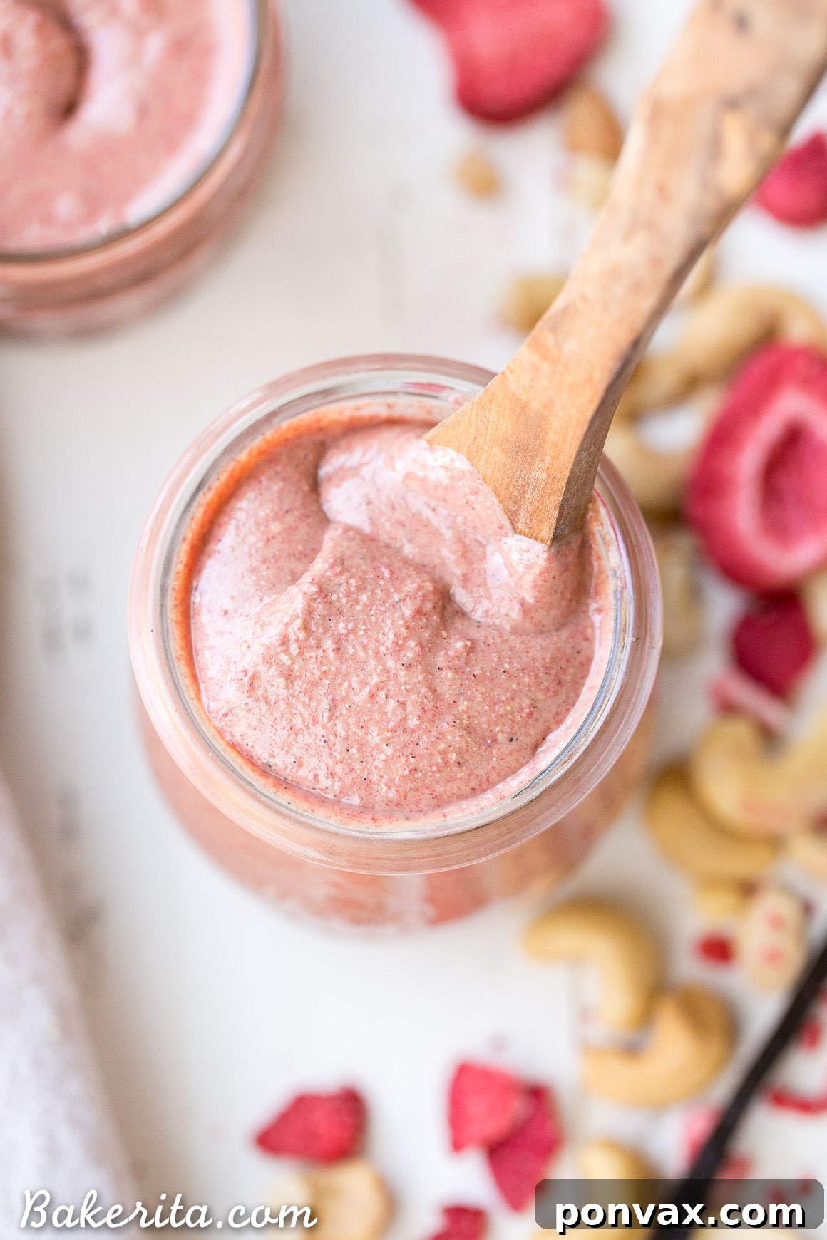 A wide shot of a glass jar filled with Strawberry Cashew Butter, with a spoon in it, surrounded by fresh strawberries.