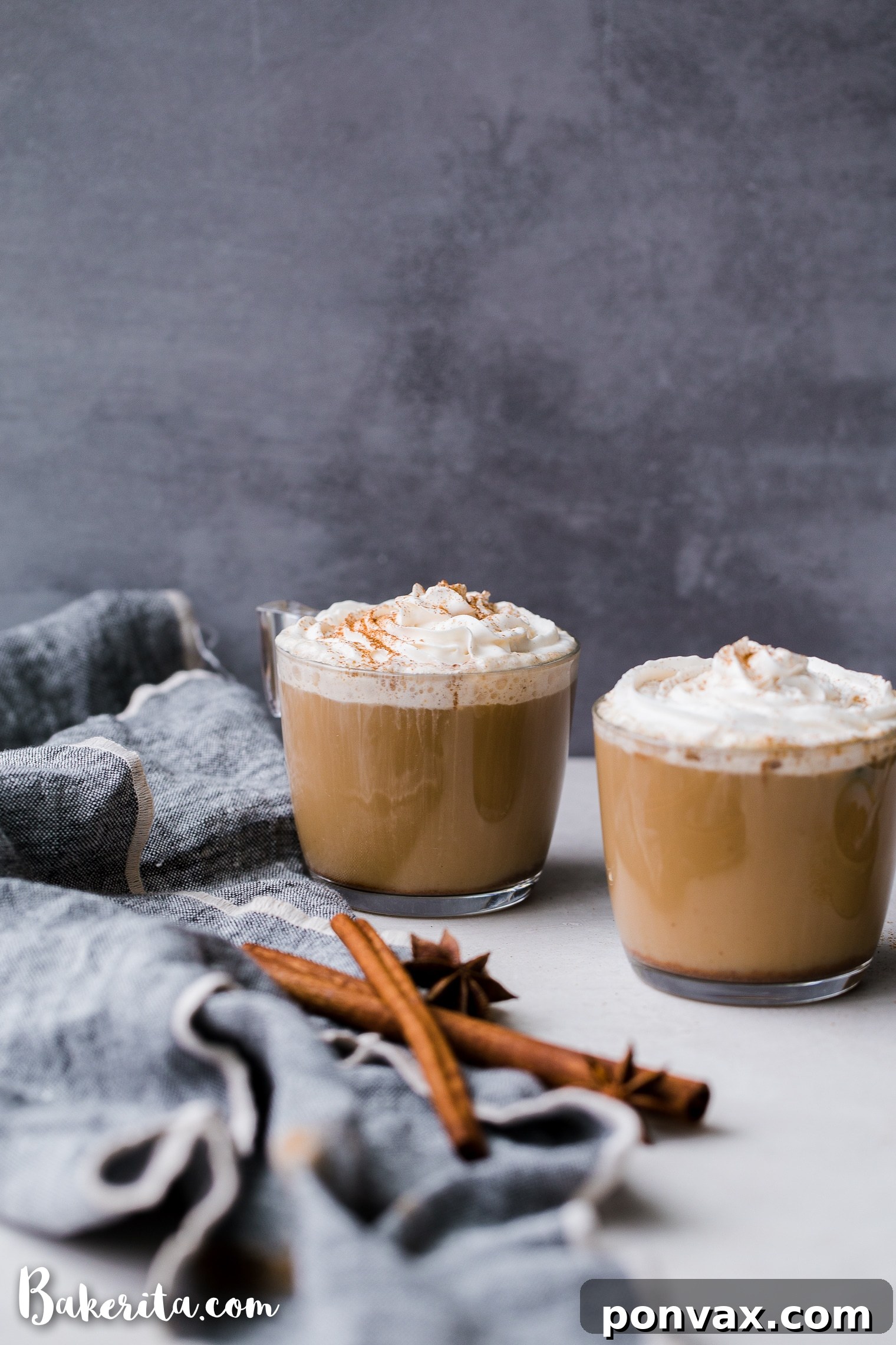 A hand holding a clear glass mug of a beautiful Vegan Chai Latte, showcasing the beverage's rich color and a gentle sprinkle of spice on top, implying enjoyment and warmth.