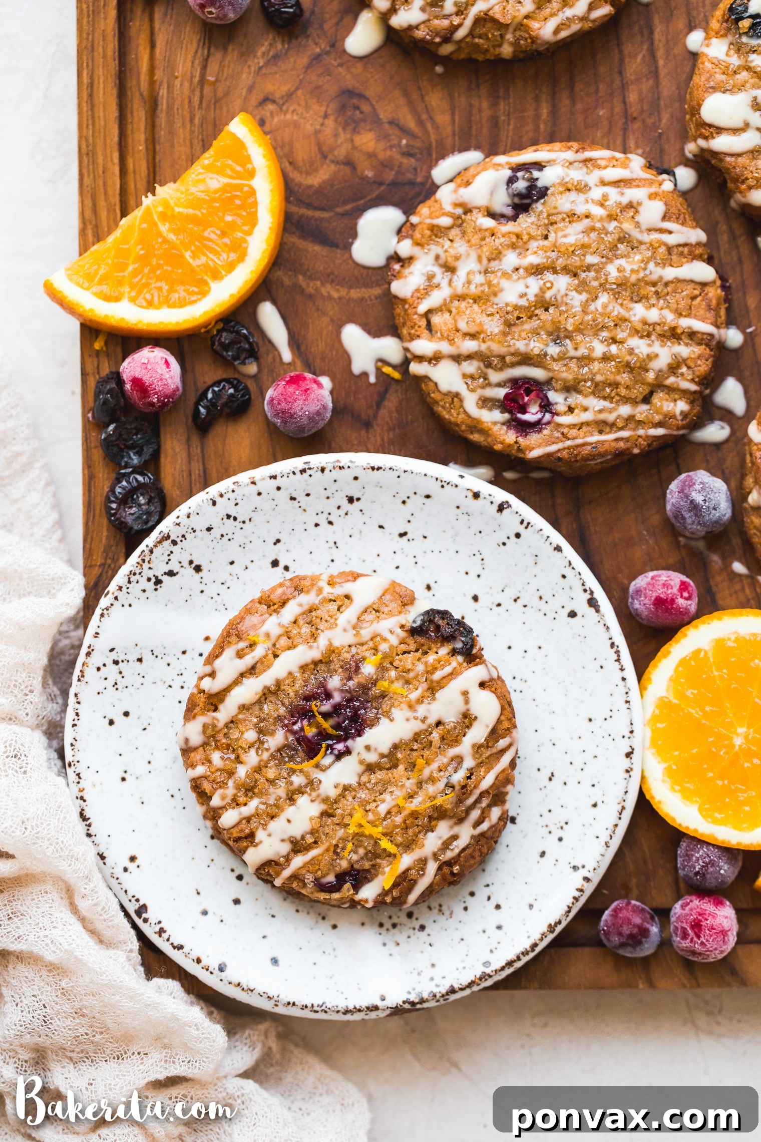 A beautifully glazed gluten-free vegan cranberry orange scone, with fresh cranberries and orange slices in the background.