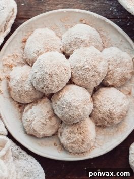 A plate of round snowball cookies covered in powdered sugar sits on a wooden surface, surrounded by a white textured cloth. The cookies are arranged closely together, creating a cozy and inviting presentation.