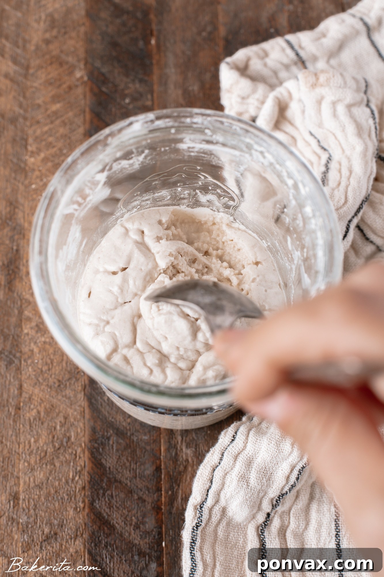 A hand mixing gluten-free flour and water in a glass jar to start a sourdough starter, with a rubber band marking the initial level.