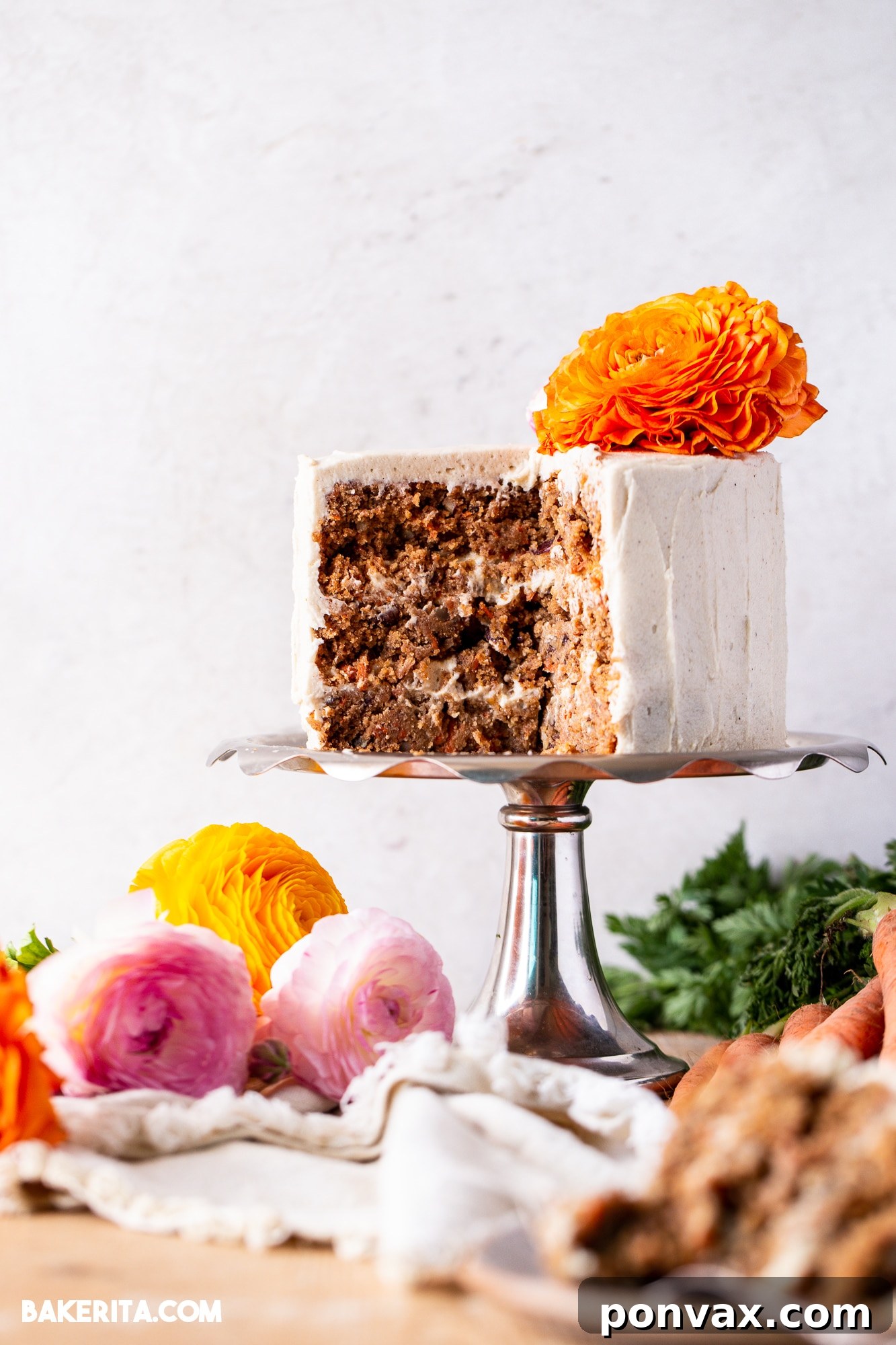 A three-layer gluten-free vegan carrot cake adorned with a delicate white cream cheese frosting, surrounded by fresh flowers.