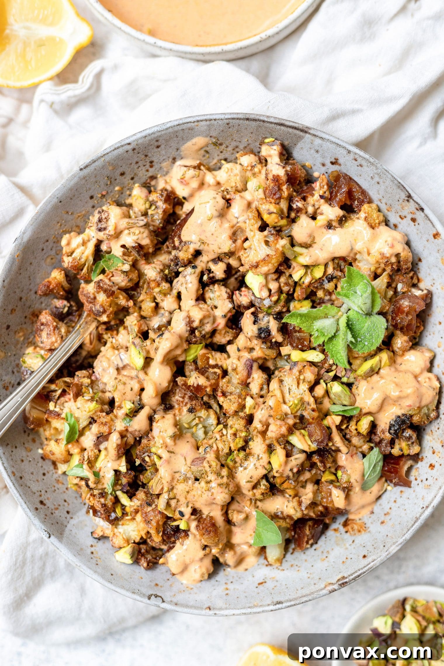 Golden Charred Cauliflower with Zesty Tahini Drizzle 7 Top down close-up photo of tahini cauliflower in a white speckled pottery bowl. Cauliflower is drizzled with a tahini harissa sauce and topped with fresh mint sprigs. A spoon is present on the left of the bowl.