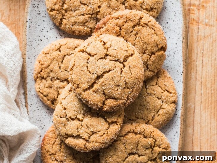 25+ Delicious Vegan Cookie Recipes 2 A speckled rectangular plate holds a pile of cracked, sugar-coated vegan sugar cookies on a wooden surface, with a light cloth and a bowl of sugar nearby.
