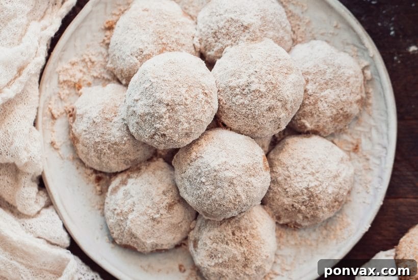 25+ Delicious Vegan Cookie Recipes 7 A pile of round, powdery white snowball cookies on a white plate, garnished with a few green leaves and red berries, creating a festive winter scene.