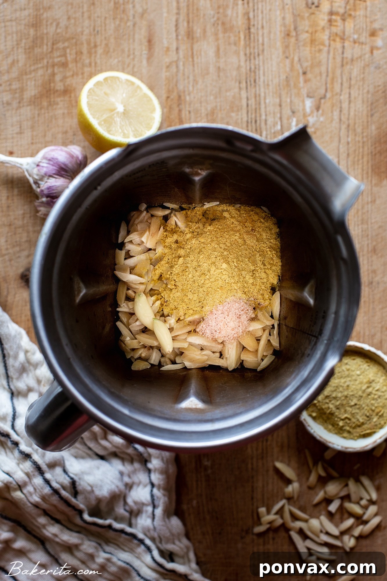 Velvety 5-Minute Vegan Almond Ricotta 4 A robust metal Vitamix blender container, brimming with the raw ingredients destined to become creamy vegan ricotta. Inside, slivered almonds are visible alongside nutritional yeast, salt, wedges of fresh lemon, and cloves of garlic, poised for blending into a smooth, plant-based cheese.