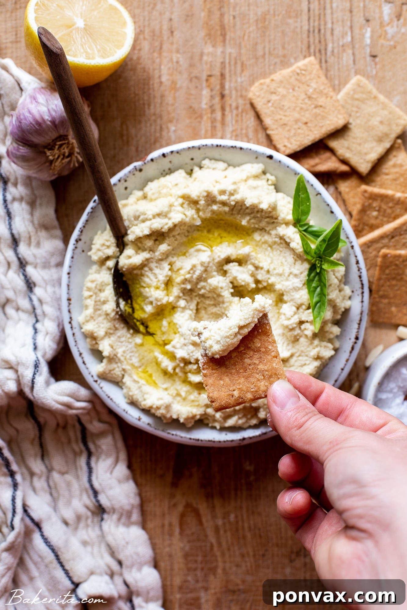 Velvety 5-Minute Vegan Almond Ricotta 6 A white ceramic bowl filled with vibrant vegan ricotta, adorned with fresh basil leaves and a delicate drizzle of olive oil. A hand holds a cracker, generously spread with the creamy vegan ricotta, poised above the bowl for a delightful bite. This image captures the delicious appeal and versatility of the plant-based cheese.
