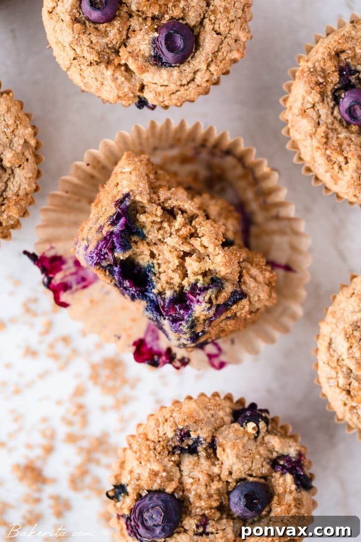 A blueberry muffin lying on its side, with a bite taken out to reveal the moist, blueberry-studded interior, nestled in a parchment wrapper amidst other muffins.