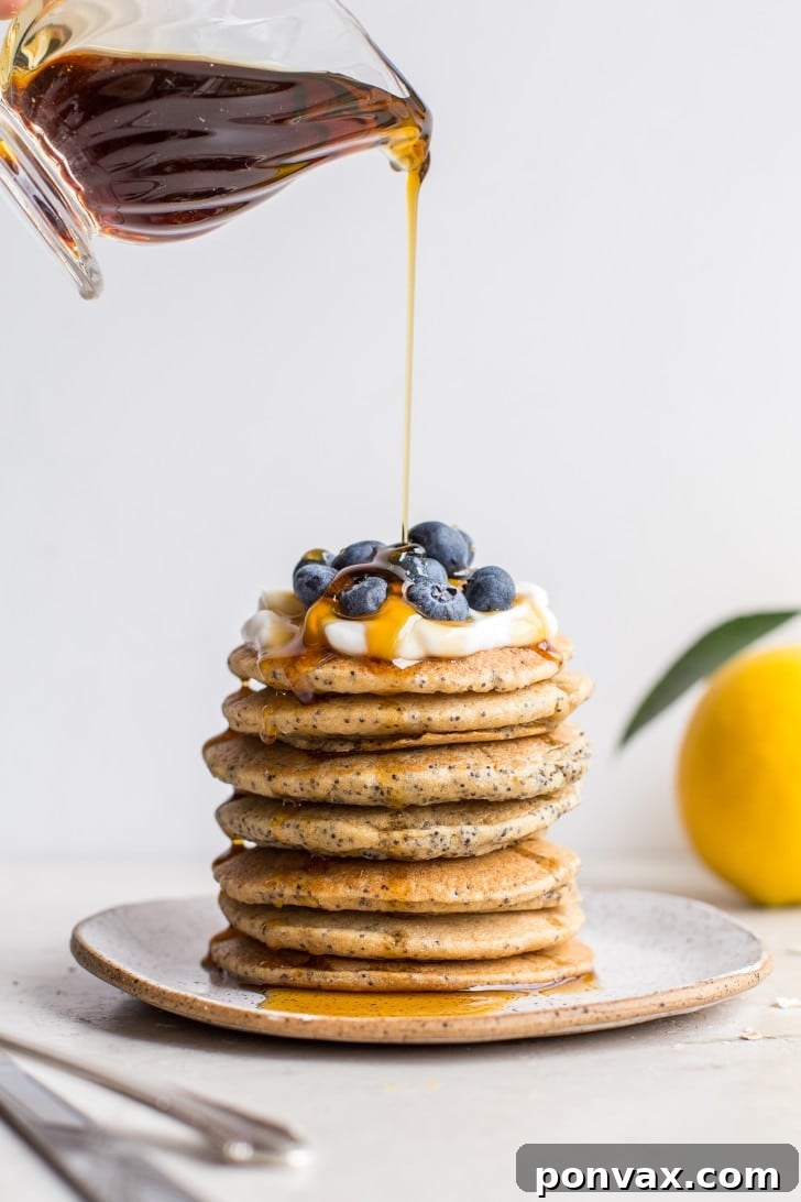 Fluffy stack of Vegan Lemon Poppyseed Pancakes drizzled with maple syrup and fresh lemon slices.