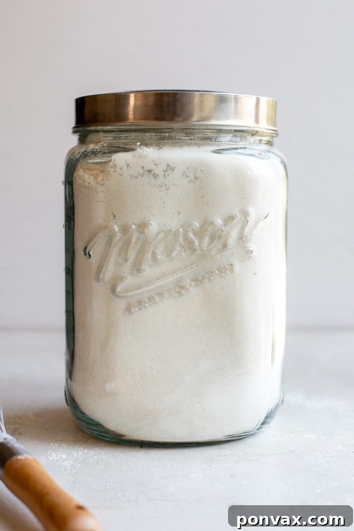 A large mason jar filled with a homemade Gluten-Free Bread Flour Blend, positioned on a clean white background. A whisk rests in the foreground, ready for use, emphasizing the DIY nature of the blend.