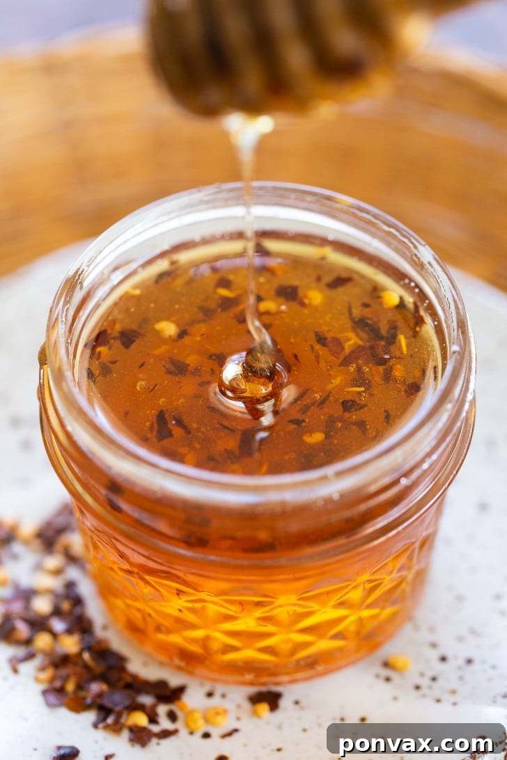 A mason jar filled with homemade hot honey, showing visible chili flakes suspended in the golden liquid, placed on a white plate.