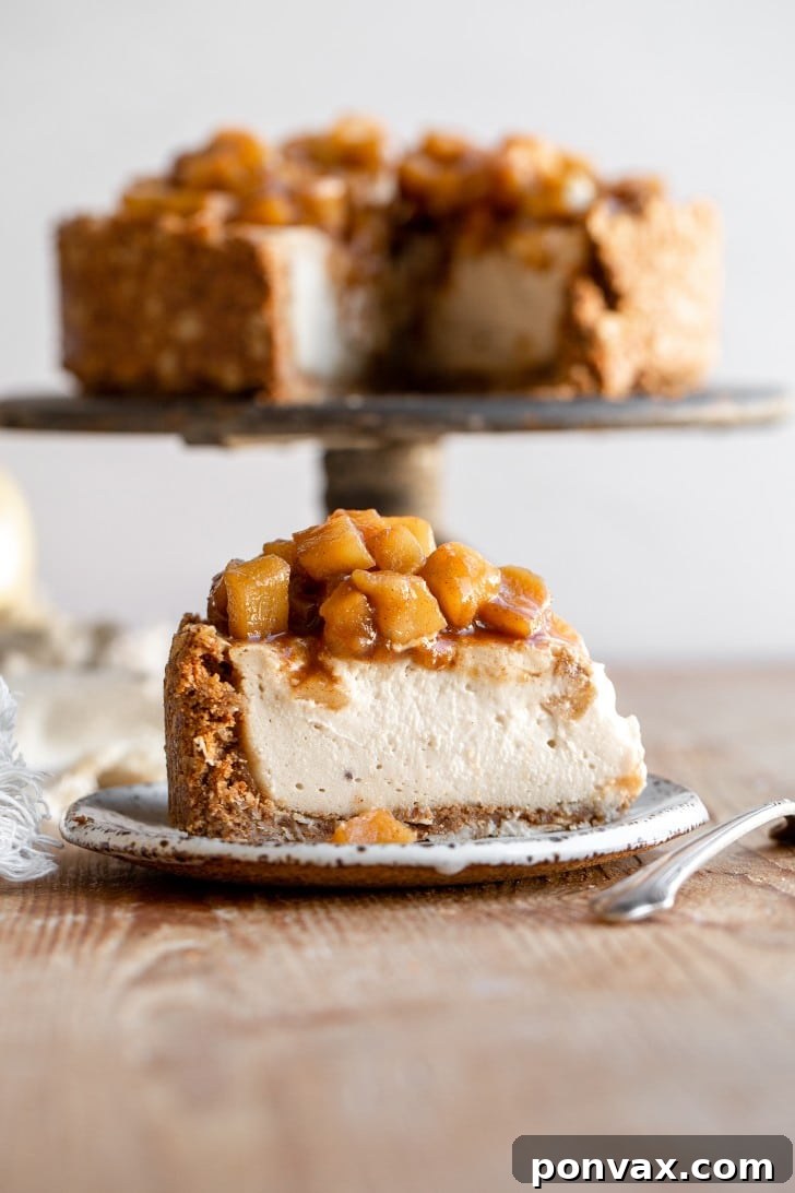 A beautifully presented slice of vegan baked apple pie cheesecake on a white plate, positioned on a rustic wooden board. The rest of the whole cheesecake, adorned with a golden apple topping, sits on a cake stand in the background, creating an inviting scene.