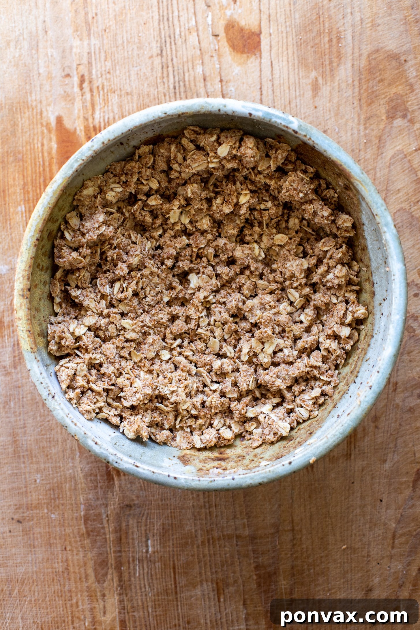 Close-up shot of the raw, mixed ingredients for the gluten-free oatmeal crust in a mixing bowl, ready to be pressed into a pan. The texture of the oats and almond flour is visible, indicating a wholesome base for the vegan apple cheesecake.