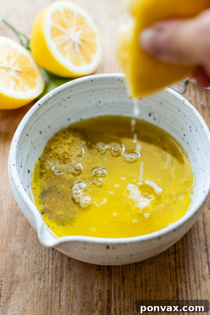 A whisk vigorously mixing lemon juice, garlic, and Dijon mustard in a bowl, demonstrating the emulsification process.