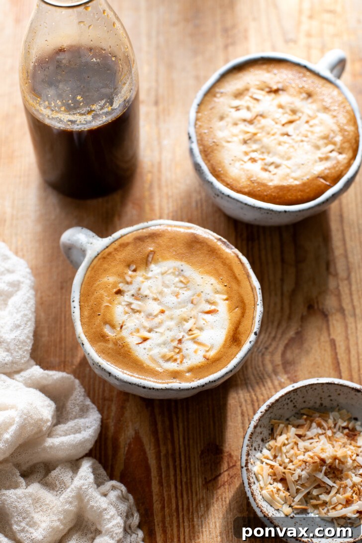 Tropical Coconut Elixir 2 A glass of homemade coconut latte topped with froth, garnished with a sprinkle of coconut flakes, with a bottle of homemade coconut syrup in the background.