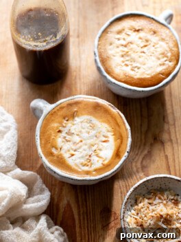 A glass of homemade coconut latte topped with froth, garnished with a sprinkle of coconut flakes, with a bottle of homemade coconut syrup in the background.