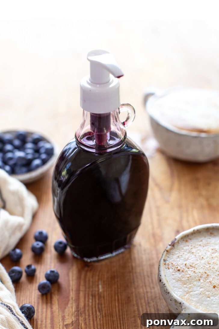 Sweet Blueberry Elixir: Elevate Your Lattes, Cocktails, and Culinary Creations 2 A glass bottle of homemade blueberry syrup sits on a wooden board. In front, a blueberry oat milk latte in a ceramic mug, and a small bowl of fresh blueberries complete the serene rustic scene.
