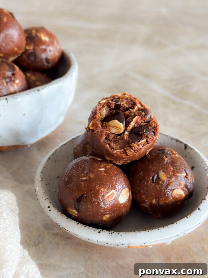 A perfectly portioned chocolate oatmeal bite with a small bite taken out, resting atop two other delicious oatmeal treats in a small white ceramic bowl. In the background, another bowl brimming with these energy bites sits on a cream marble countertop, inviting you to grab one.