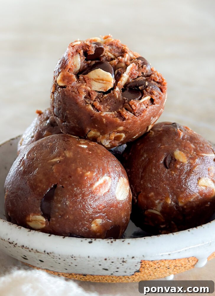 A perfectly formed chocolate oatmeal ball with a small bite taken out, resting delicately atop two other delightful energy bites in a charming small white ceramic bowl. This close-up highlights the rich texture and delicious appeal of the snack.