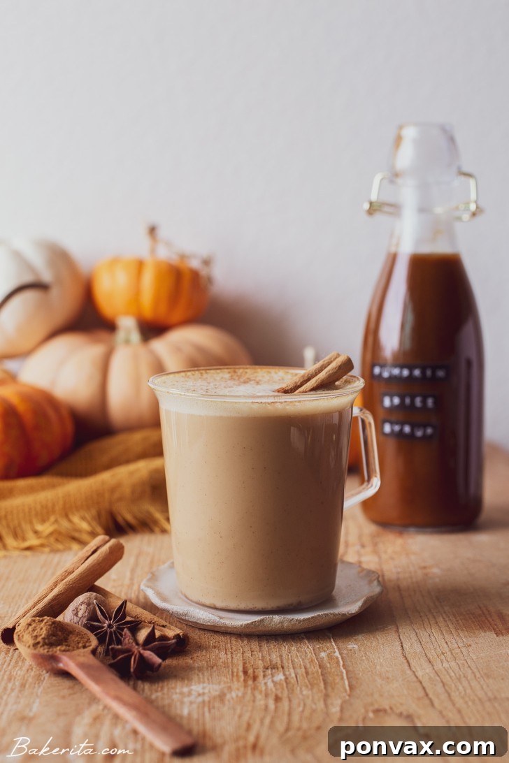 A beautifully styled pumpkin spice latte in a clear mug, garnished with cinnamon and a sprig of rosemary, sitting on a wooden surface next to fresh pumpkins and fall leaves.