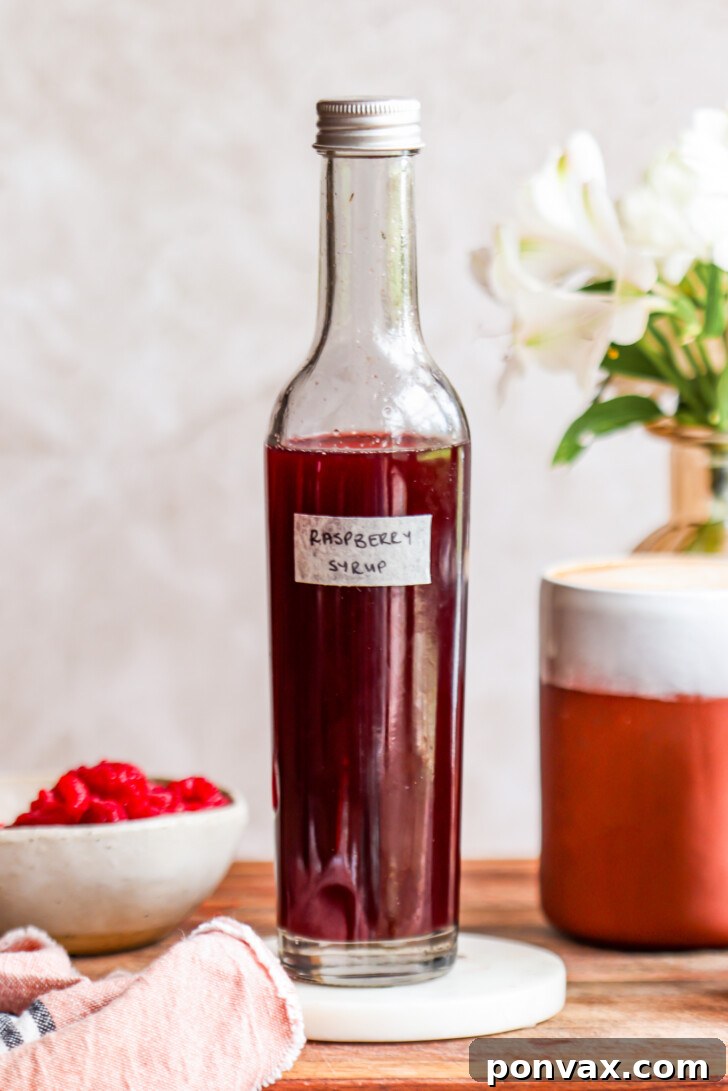 A vibrant, crimson red homemade raspberry syrup poured into a clear glass bottle, surrounded by fresh raspberries, suggesting its natural and fresh ingredients. Perfect for coffee, cocktails, or breakfast.