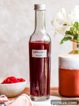 A vibrant, crimson red homemade raspberry syrup poured into a clear glass bottle, surrounded by fresh raspberries, suggesting its natural and fresh ingredients. Perfect for coffee, cocktails, or breakfast.