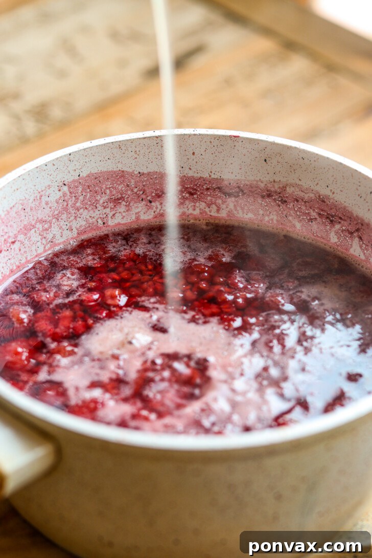 Close-up of the raspberries fully broken down and deeply colored after simmering, with a spoon stirring the rich, liquid mixture.