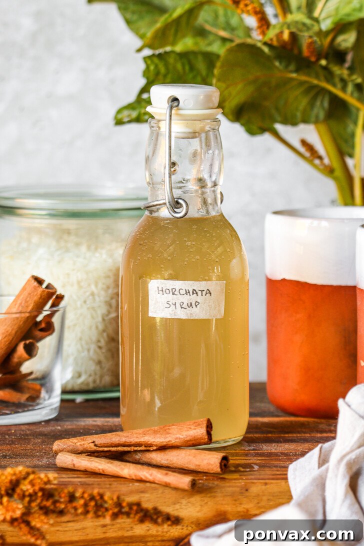 A glass swing-top bottle labeled 'Horchata Syrup' filled with light golden syrup, surrounded by cinnamon sticks, a jar of uncooked white rice, and ceramic mugs on a wooden surface with green leaves in the background.