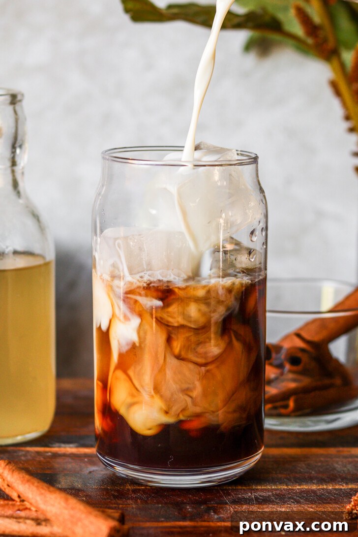 DIY Horchata Oatmilk Shaken Espresso Starbucks Style 2 Iced coffee with horchata syrup and oat milk swirling in a glass, with a bottle of homemade horchata syrup and cinnamon sticks on a wooden surface.