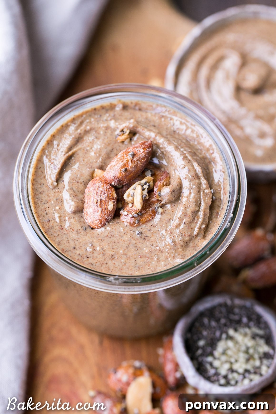A jar of Maple Almond Butter with a spoon, surrounded by loose almonds and seeds.